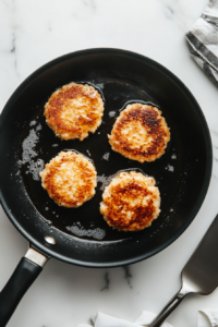 This image shows crab cakes cooking in a skillet, their surfaces turning golden and crispy as they sizzle in a layer of hot oil.