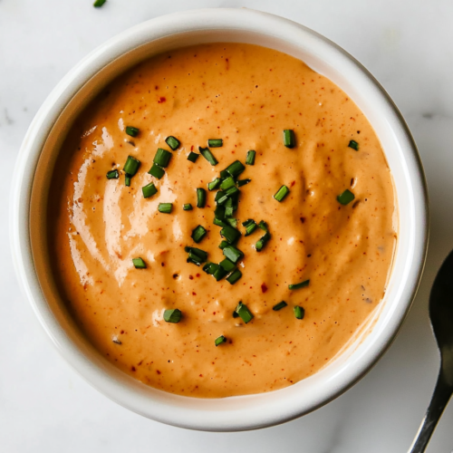 This image shows a rich, creamy crab cake sauce with a vibrant orange hue, made from mayonnaise, mustard, lemon juice, hot sauce, Creole seasoning, and other spices, elegantly topped with finely chopped green onions.