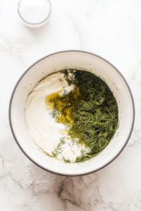 This image shows a person using paper towels to gently pat dry cucumber and onion slices, ensuring they absorb the creamy sour cream dressing more effectively.