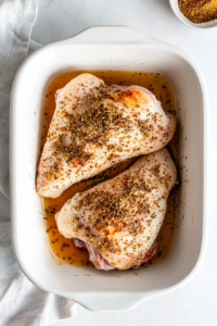 This image shows split chicken breast marinating in a baking dish with herbs, spices, and seasonings, allowing the flavors to deeply infuse before baking.