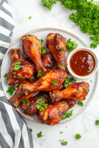 This image shows perfectly glazed Lollipop Chicken Legs arranged on a plate, served with a side of extra BBQ sauce for dipping.