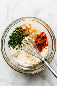 This image shows a whisk blending mayonnaise, mustard, hot sauce, and spices in a mixing bowl, creating a smooth, creamy, and flavorful sauce perfect for pairing with crab cakes.