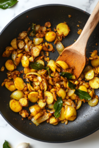 This image shows a sizzling mixture of finely chopped ginger, minced garlic, and fresh curry leaves cooking in hot oil in a black frying pan, releasing their rich, aromatic flavors.